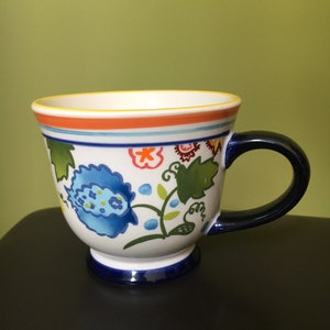 May include: A white ceramic mug with a blue, green, orange, and yellow floral design. The mug has a blue handle and a blue rim.