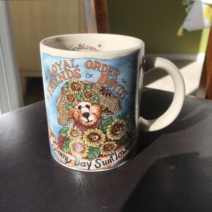 May include: A white ceramic mug with a blue and yellow design featuring a teddy bear wearing a sunflower hat and the text "The Loyal Order of Boyds Sunny Day Sunflowers".
