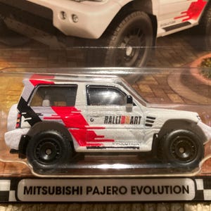 May include: A white Mitsubishi Pajero Evolution diecast model car with black wheels and red and black graphics. The words "Ralli Art" and "Evolution" are visible on the side. The car is in a clear plastic package.