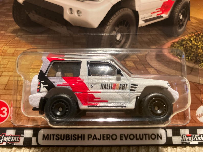 May include: A white Mitsubishi Pajero Evolution diecast model car with black wheels and red and black graphics. The words "Ralli Art" and "Evolution" are visible on the side. The car is in a clear plastic package.