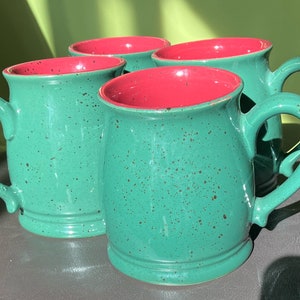 May include: Four green ceramic mugs with red interiors. The mugs have a speckled glaze and are perfect for a hot beverage.