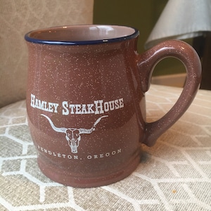 May include: A brown speckled ceramic mug with a blue rim. The mug has a white logo that says "Hamley Steakhouse" with a stylized steer skull and the text "Pendleton, Oregon".