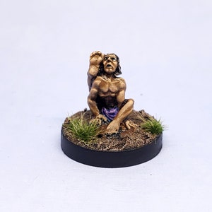 Puede incluir: Una figurita miniatura pintada de una persona sentada en una postura de yoga con la pierna levantada detrás de la cabeza. La figurita está pintada en tonos marrones y tiene un paño morado alrededor de la cintura. La figurita está sentada sobre una base redonda con tierra marrón y hierba verde.