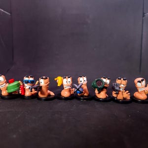 Puede incluir: Un conjunto de ocho pequeñas figuras de gusanos de color tostado, estilo dibujos animados, sobre bases circulares negras individuales. Cada gusano tiene una expresión facial única y sostiene un accesorio diferente, incluyendo una mina verde, un plátano y un bate de béisbol.