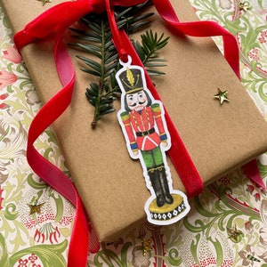 Op de afbeelding: Een cadeau verpakt in bruin papier, versierd met een rode fluwelen lint en een notenkrakerlabel. Het cadeau is versierd met groen, rode bessen en gouden sterconfetti, wat een feestelijk thema creëert.