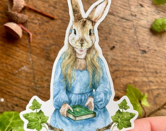 May include: A sticker featuring a watercolor illustration of a rabbit wearing a blue dress and holding a green book. The rabbit is surrounded by green ivy.