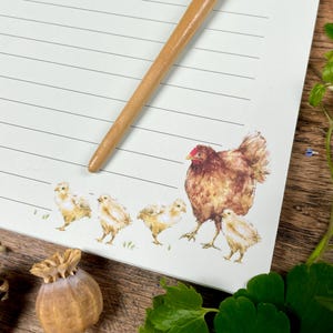 Op de afbeelding: Een gelijnd notitieblok met een aquarel illustratie van een kip en kuikens. Een houten penhouder ligt op het papier. De kip is bruin en de kuikens zijn geel. Het notitieblok ligt op een houten oppervlak.