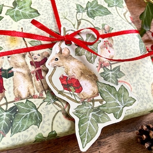 May include: A rectangular gift box wrapped in light green paper featuring a pattern of green ivy leaves and mice. A bright red ribbon is tied around the box, securing a die-cut gift tag shaped like a mouse holding a red gift box among ivy leaves.