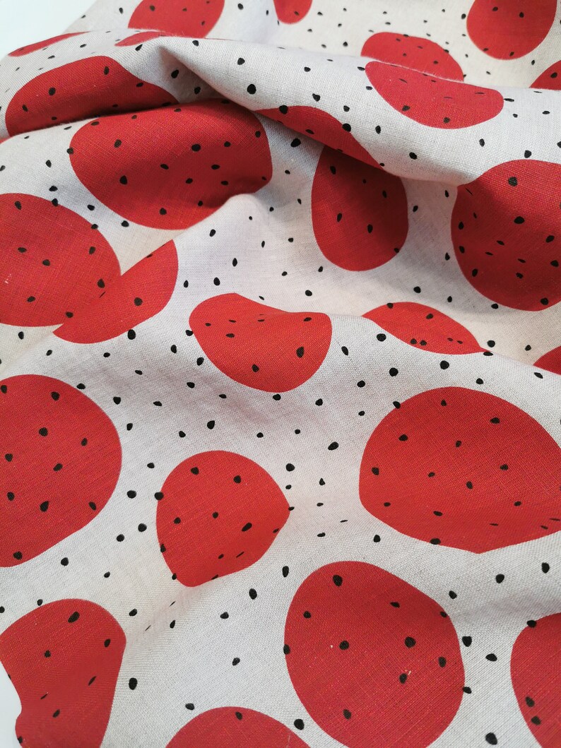 May include: White fabric with a repeating pattern of red circles with black dots. The fabric is folded and wrinkled.