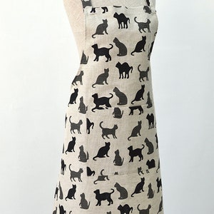 May include: A beige linen apron with a black cat silhouette pattern. The apron has two pockets and adjustable straps.