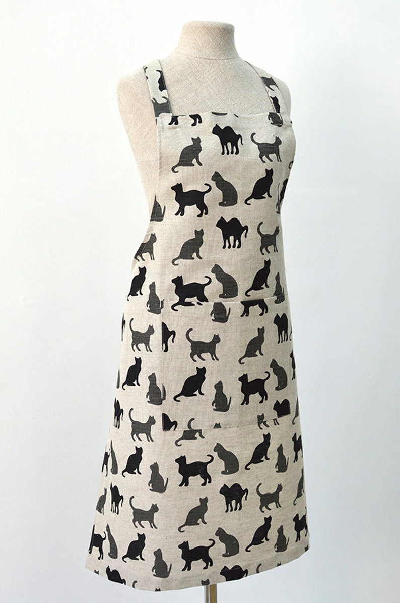 May include: A beige linen apron with a black cat silhouette pattern. The apron has two pockets and adjustable straps.