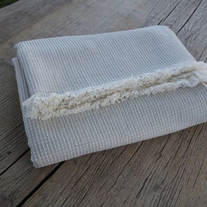 May include: A folded, light grey throw blanket with a fringe border. The blanket is made of a soft, textured fabric.