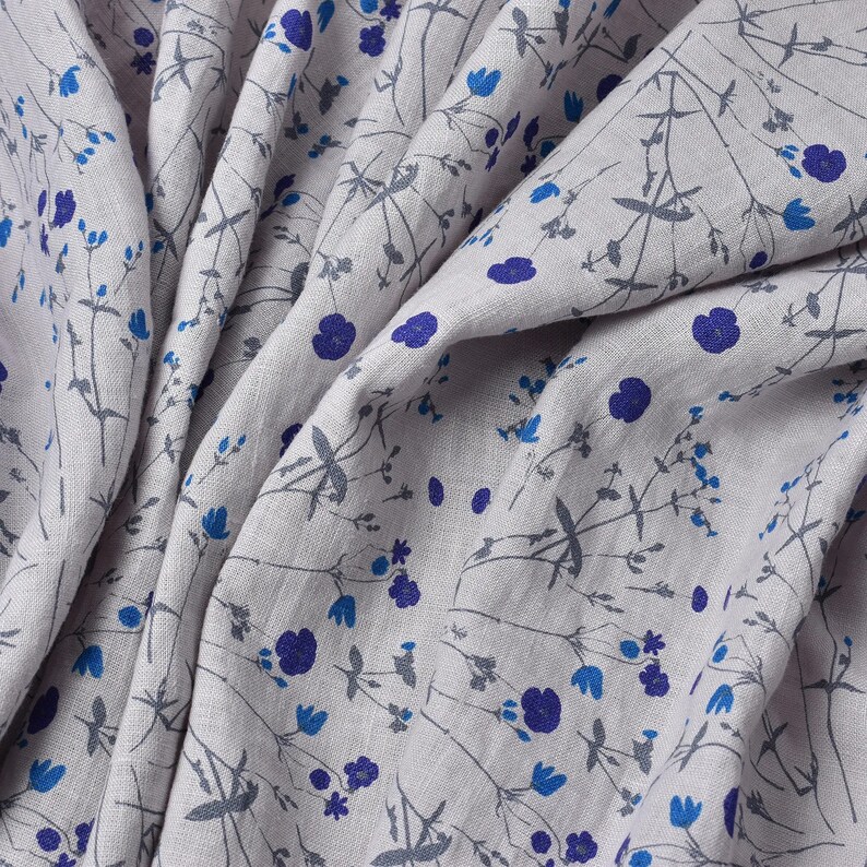 Puede incluir: Un primer plano de una tela de lino gris claro con un patr&oacute;n repetitivo de peque&ntilde;as flores azules y tallos grises. La tela est&aacute; doblada y drapeada, mostrando la textura y el detalle del dise&ntilde;o.