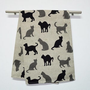 May include: A beige kitchen towel with a black silhouette pattern of cats. The cats are in various poses, including sitting, standing, and lying down.