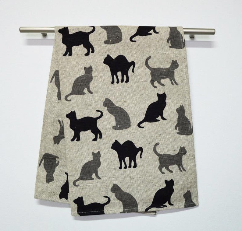 May include: A beige kitchen towel with a black silhouette pattern of cats. The cats are in various poses, including sitting, standing, and lying down.