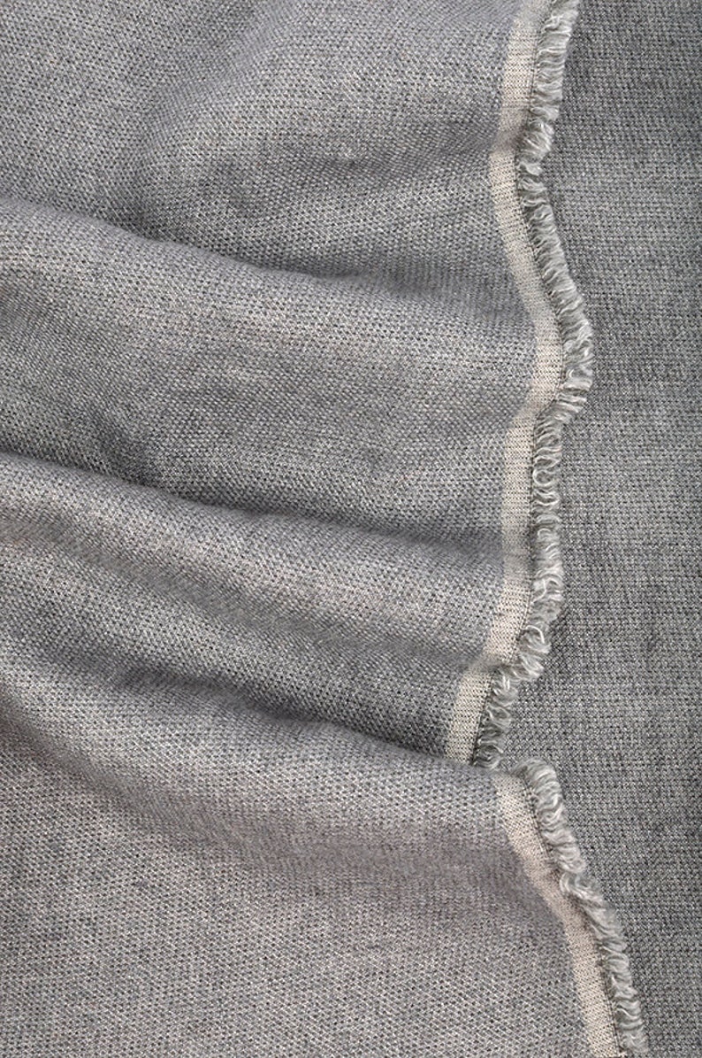 May include: Close-up of a gray fabric with a textured weave. The fabric has a frayed edge with a thin, off-white stripe. The material is folded, showing the texture and drape of the textile.