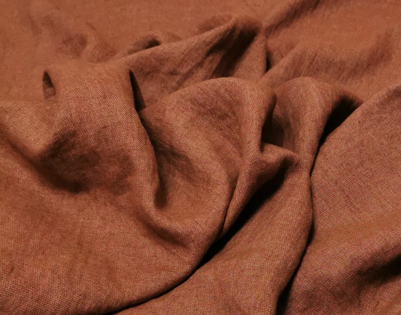 Puede incluir: Primer plano de una tela de lino marr&oacute;n con un tejido texturizado. La tela est&aacute; doblada y drapeada, mostrando su textura suave y natural.
