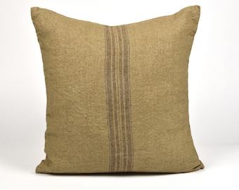 May include: Square pillow with a textured, tan fabric cover. A vertical stripe of three brown lines runs down the center. The pillow has a soft, casual appearance, suitable for home decor.