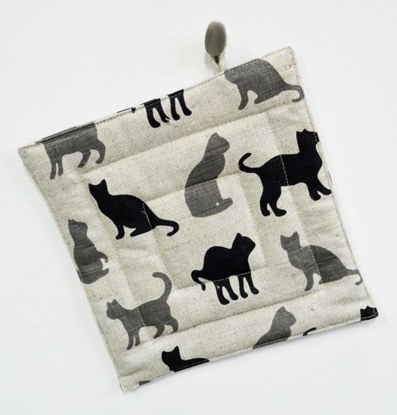 May include: A beige quilted pot holder with a black cat pattern. The pot holder has a loop for hanging.