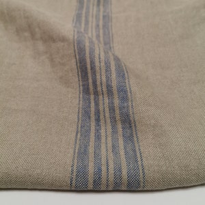 K&ouml;nnte beinhalten: Ein Nahaufnahme von einem beigefarbenen Leinenstoff mit einem blau-wei&szlig; gestreiften Muster. Der Stoff ist in der Mitte gefaltet, so dass das gestreifte Muster auf der einen Seite und der einfarbige beige Stoff auf der anderen Seite zu sehen ist.