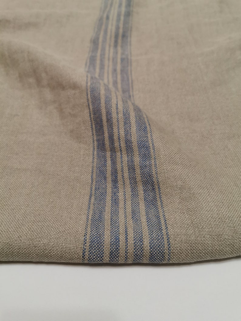 K&ouml;nnte beinhalten: Ein Nahaufnahme von einem beigefarbenen Leinenstoff mit einem blau-wei&szlig; gestreiften Muster. Der Stoff ist in der Mitte gefaltet, so dass das gestreifte Muster auf der einen Seite und der einfarbige beige Stoff auf der anderen Seite zu sehen ist.