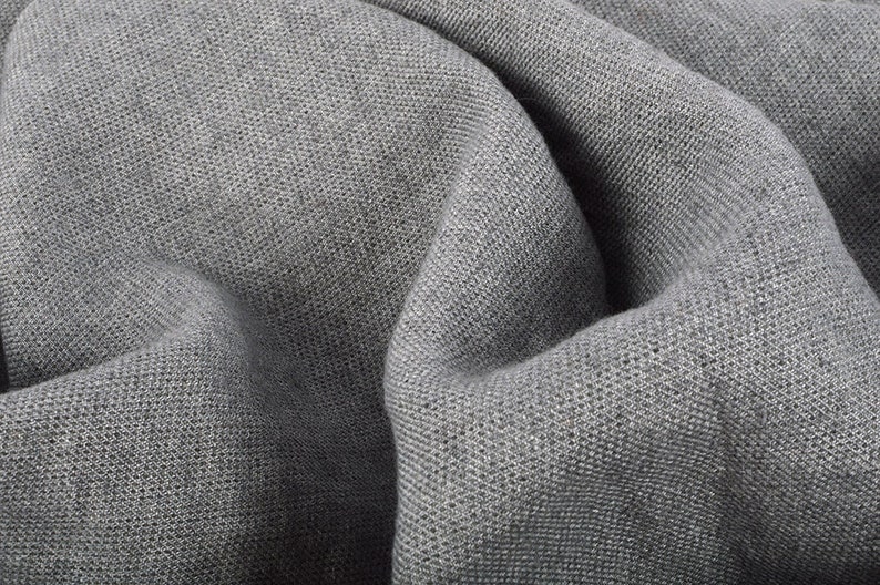 May include: Close-up of a gray fabric with a textured weave. The fabric is draped, creating soft folds and shadows. The material appears to be a medium-weight textile, suitable for clothing or home decor.