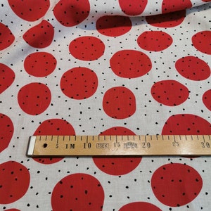 May include: White fabric with a red polka dot pattern. The dots are large and have a black speckling pattern. A wooden ruler with black markings is placed on the fabric. The ruler is marked with centimeters and inches.