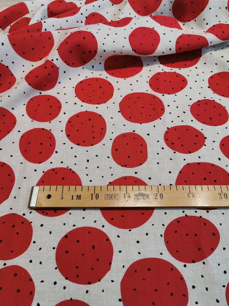 May include: White fabric with a red polka dot pattern. The dots are large and have a black speckling pattern. A wooden ruler with black markings is placed on the fabric. The ruler is marked with centimeters and inches.