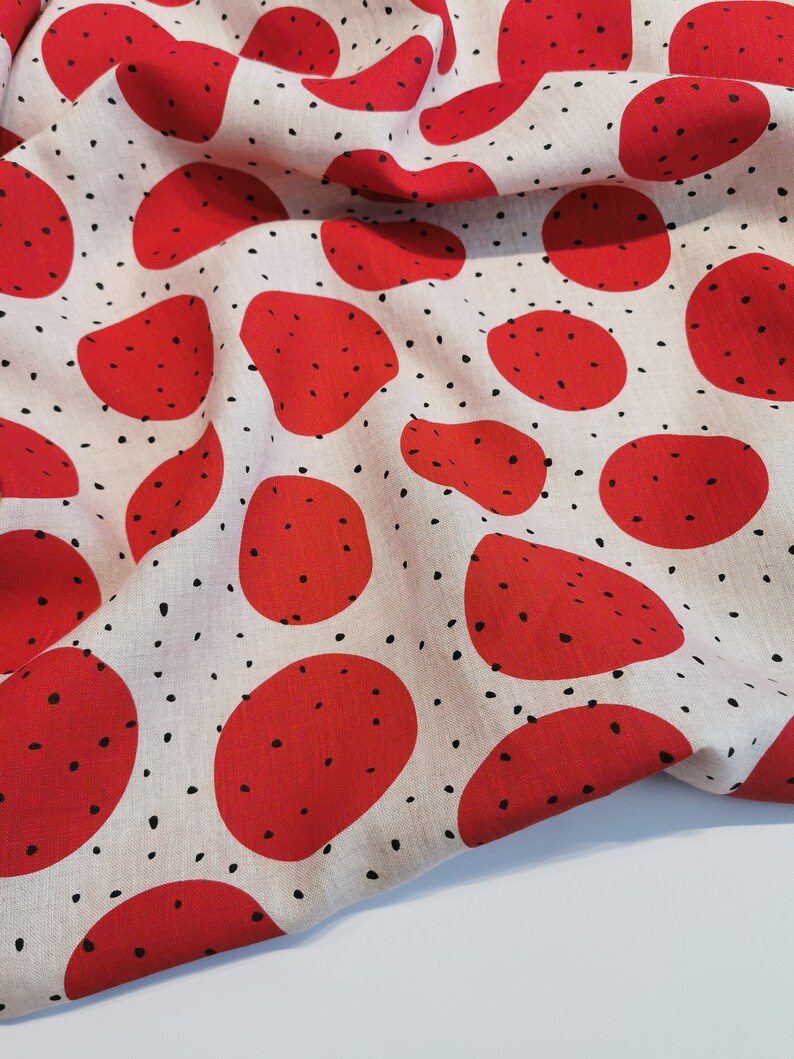 May include: Red and white fabric with a repeating pattern of red circles with black speckles. The fabric is folded and draped over a white surface.