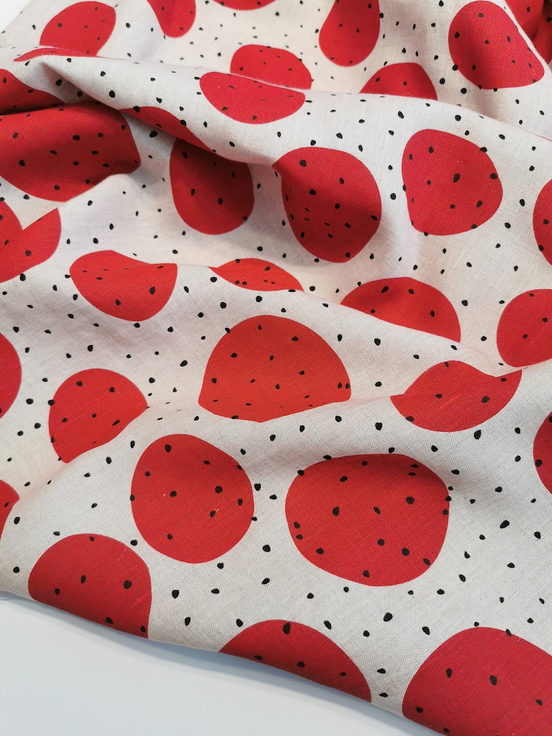 May include: Red fabric with a white background and a repeating pattern of red circles with black dots. The fabric is folded and has a textured surface.