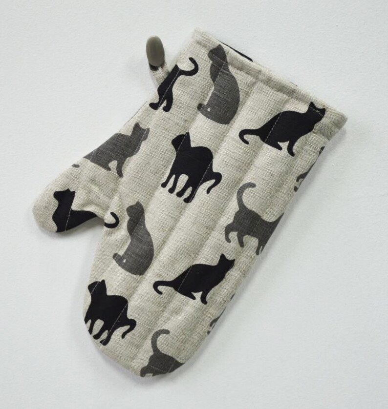 May include: A beige oven mitt with a black cat pattern. The mitt has a loop for hanging.