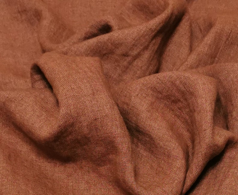 Puede incluir: Primer plano de una tela de lino marr&oacute;n con un tejido texturizado. La tela est&aacute; doblada y drapeada, mostrando su textura suave y natural.