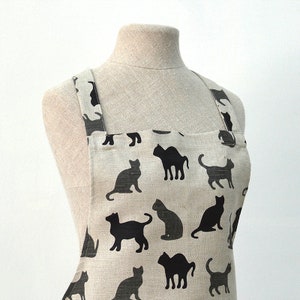 May include: A beige linen apron with black cat silhouettes printed all over. The apron has adjustable straps and a large pocket.