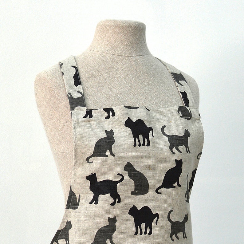 May include: A beige linen apron with black cat silhouettes printed all over. The apron has adjustable straps and a large pocket.