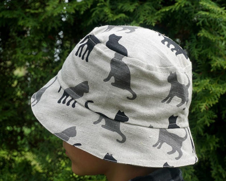 May include: A white bucket hat with a black cat print. The hat is made of a soft, lightweight fabric and has a wide brim.