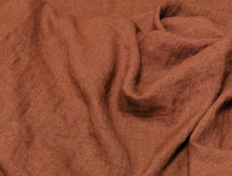 Puede incluir: Primer plano de una tela de lino de color &oacute;xido con un tejido texturizado. La tela est&aacute; doblada y drapeada, mostrando su textura suave y natural.