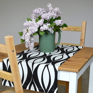 Puede incluir: Un camino de mesa con estampado en blanco y negro con un jarrón verde lleno de flores de lila sobre una mesa de madera con dos sillas de madera.