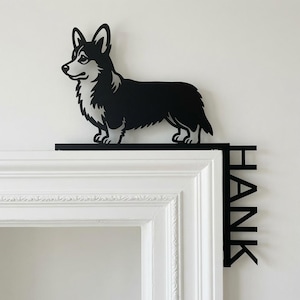 May include: A black and white metal silhouette of a Corgi dog standing on a horizontal base that extends over a door frame. The vertical side of the base features the name HANK cut out in bold, capital, black letters in a vertical orientation.