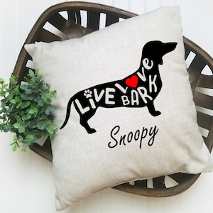 May include: A beige pillow with a black dachshund silhouette. The dachshund's body is filled with the words "Live Love Bark" and a red heart. The words "Snoopy" are written below the dachshund.