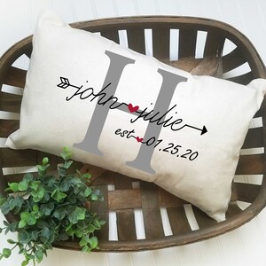 May include: A white pillow with a gray letter H, the names "john" and "julie" written in cursive, and the text "est. 01.25.20" printed on it. The pillow is in a wooden basket.