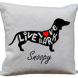May include: A white pillow with a black silhouette of a dachshund. The dachshund's body is filled with the words "Live Love Bark" and a red heart. The pillow has the name "Snoopy" written below the dachshund.