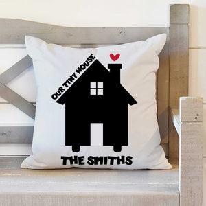 May include: White throw pillow with black silhouette of a house with a red heart on top. The text "OUR TINY HOUSE" is above the house and "THE SMITHS" is below the house.