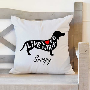 May include: White throw pillow with a black silhouette of a dachshund with the words "Live Love Bark" written on it. The heart in the word "Love" is red. The words "Snoopy" are written below the dachshund.