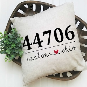May include: A beige square pillow with the text "44706 canton ohio" in black script and a red heart. The pillow is inside a brown woven basket with a green plant on the side.