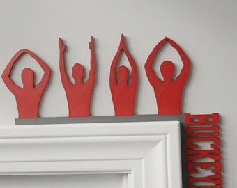 May include: A red decorative door topper featuring four silhouettes of people with arms raised in various positions, mounted on a gray L-shaped bracket. The vertical side of the bracket features the word BUCKEYES in raised red capital letters.