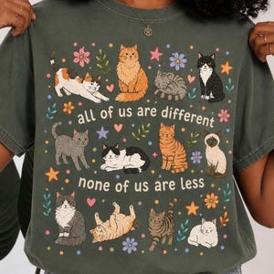 Puede incluir: Una camiseta de color verde oliva oscuro con un estampado gráfico de once gatos en varias posturas, rodeados de pequeñas flores y estrellas. El texto "all of us are different" y "none of us are less" está impreso en blanco en el centro.