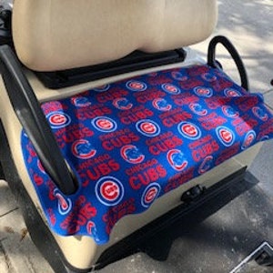 May include: A golf cart seat cover featuring a repeating pattern of the Chicago Cubs logo and the text Chicago Cubs in red and blue on a blue background. The cover is fitted over the beige bench seat of a golf cart with black armrests.