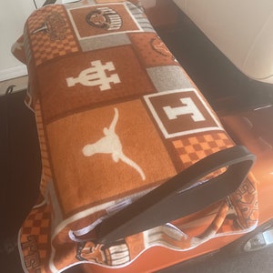 May include: A burnt orange and white blanket with various University of Texas logos and symbols. The blanket is folded and placed on a golf cart. The blanket features a Longhorn logo.