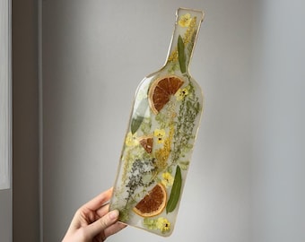 May include: A clear glass bottle shaped serving tray with a gold rim. The tray is filled with pressed flowers and dried citrus slices.