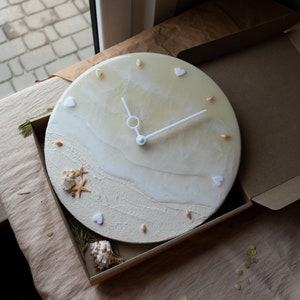 May include: A white and beige resin clock with a beach theme. The clock face has white hands and a white circle in the centre. The clock face is decorated with seashells, starfish, and white hearts. The clock is in a brown cardboard box.